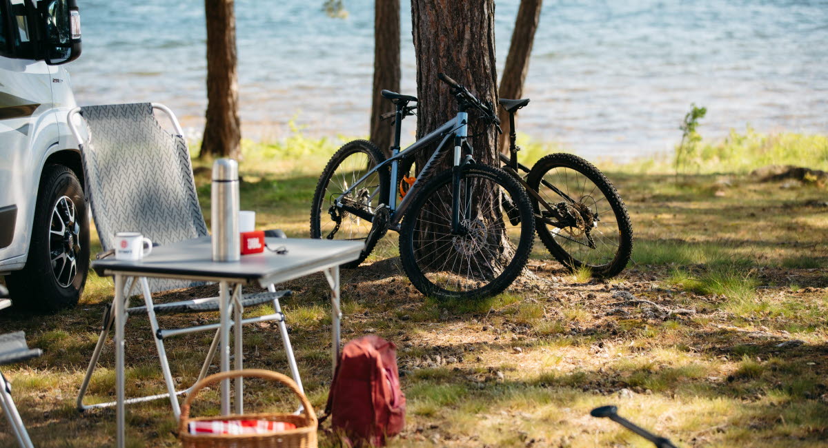Dekorativ bild föreställande en husbil, campingmöbler och en cykel vid en sjö.