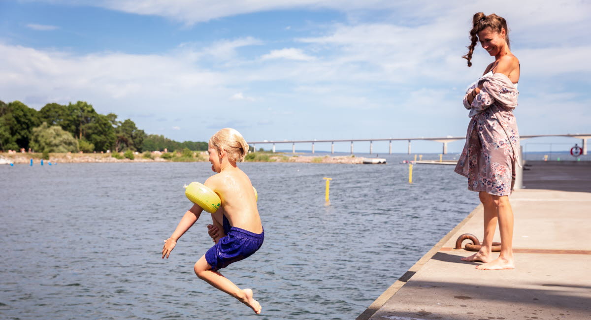 Barn hoppar i vattnet från brygga under översyn av vuxen.  