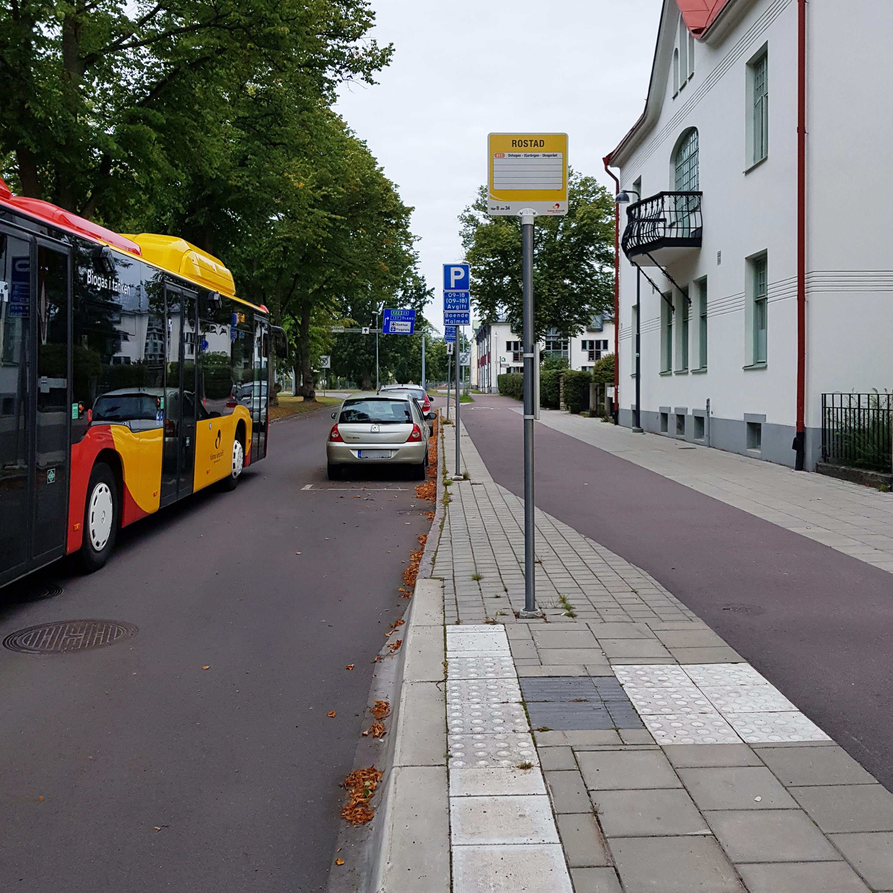 En buss och en bil på körbanan. En hållplats med höjd kant och taktila plattor samt en hållplatsskylt.