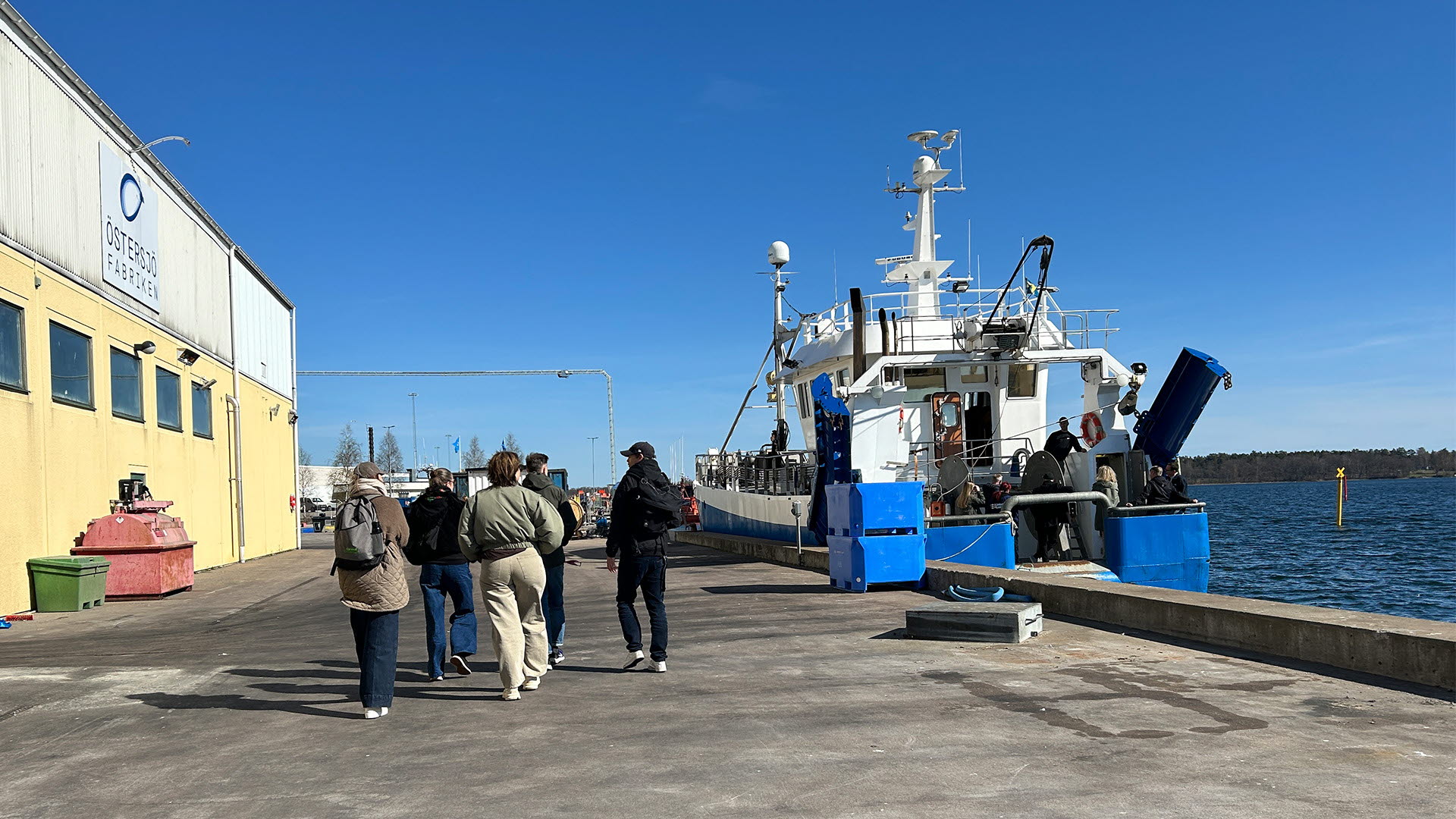 En grupp personer vid en fabrik intill en hamn med en fiskebåt.