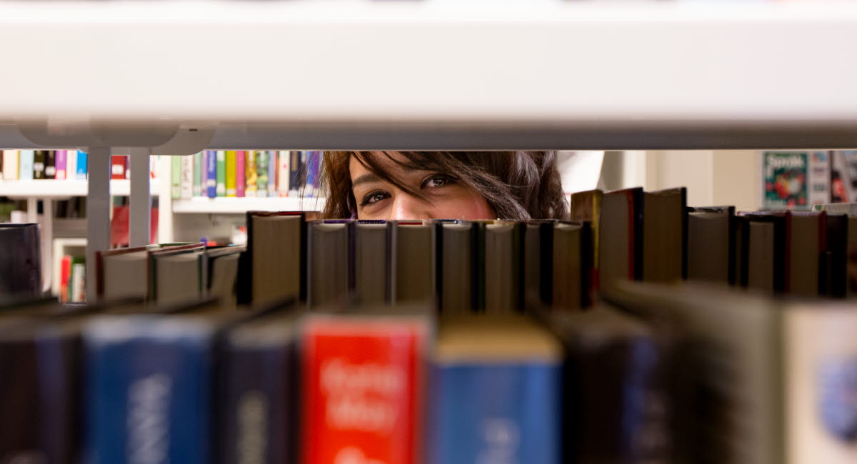 Kvinna tittar fram genom en bokhylla på ett bibliotek.